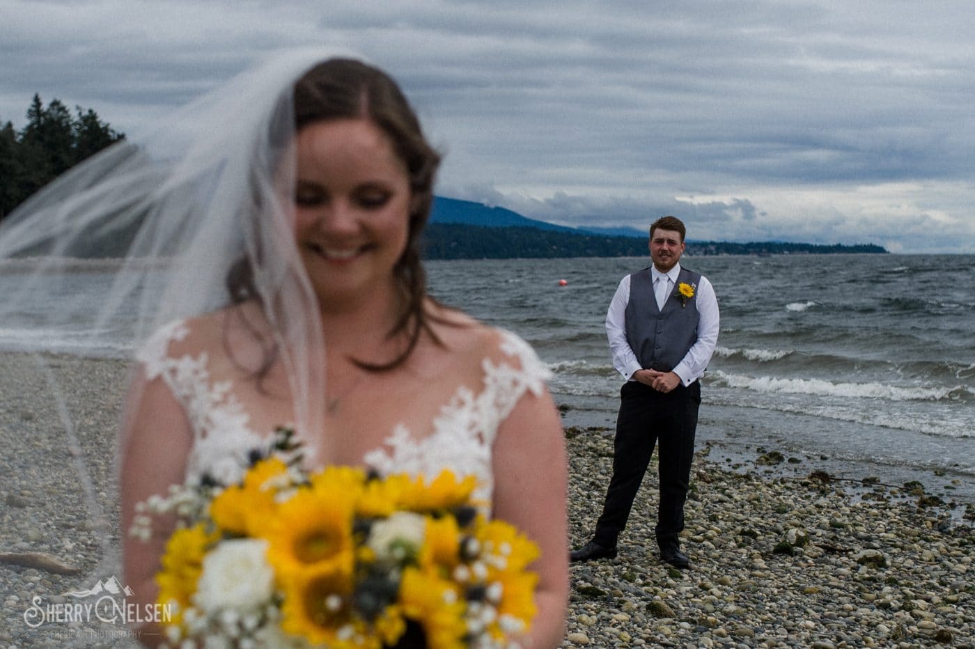groom stands behind his bride as she smiles at her gorgeous bouquet by Coastal Wedding Events