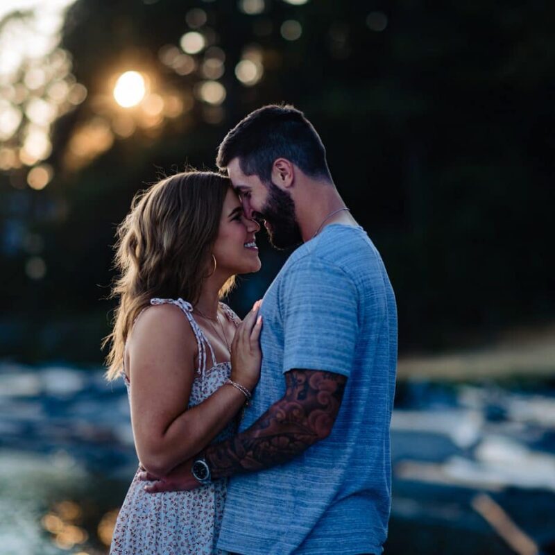 newly engaged couple enjoys the sunset as she lays her forehead against his face and they smile after their surprise proposal