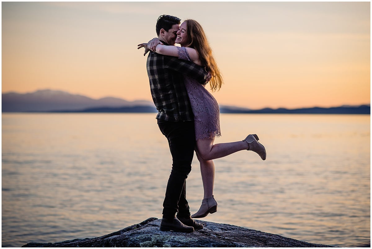 Man who has recently proposed to his love sweeps her off her feet on a rocky outcropping overlooking the ocean in Sechelt, BC.