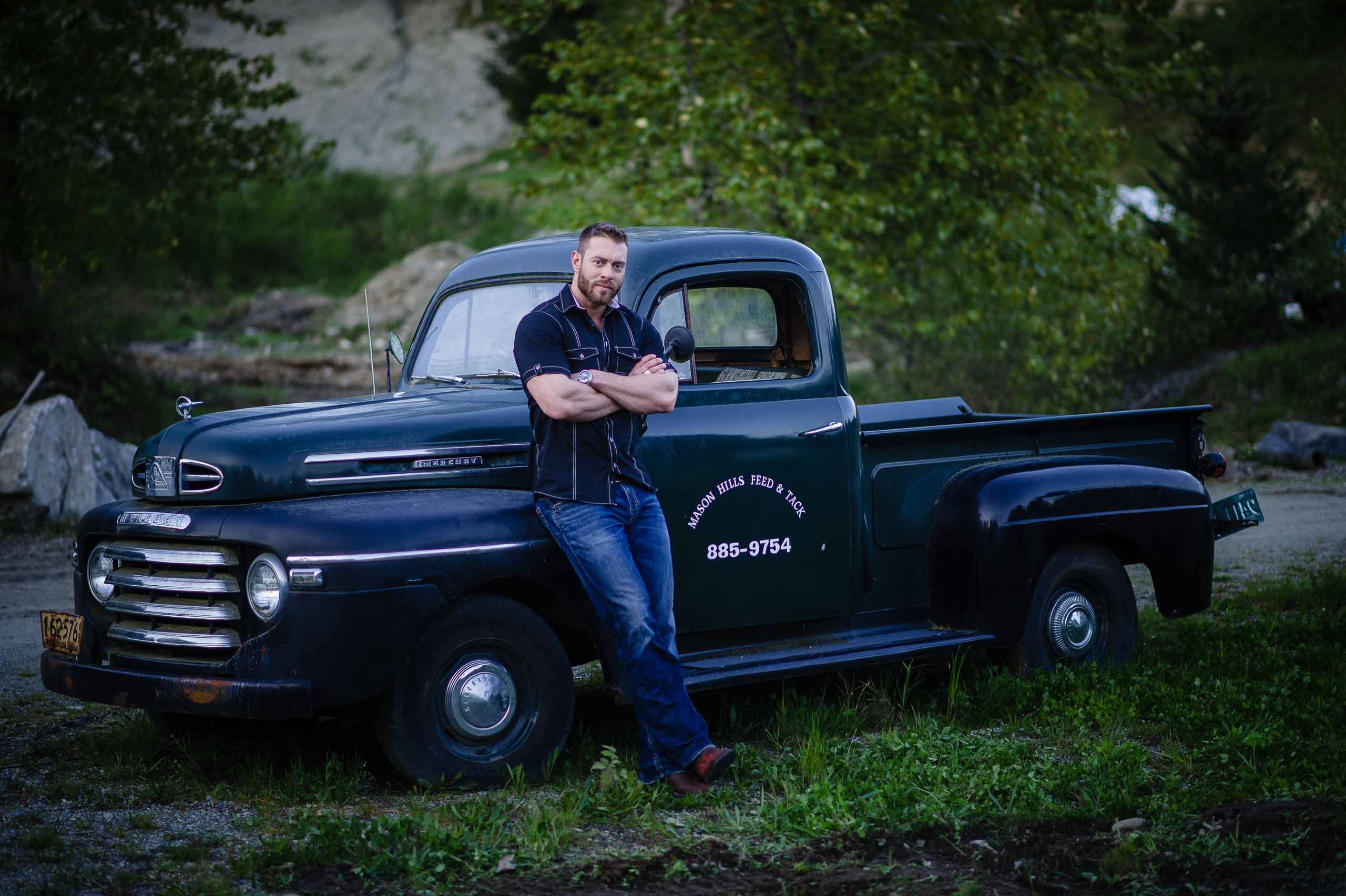 Young, good looking man wearing a dressy button up shirt and jeans leans against an old Mercury truck on a farm for photos based in Sechelt, BC
