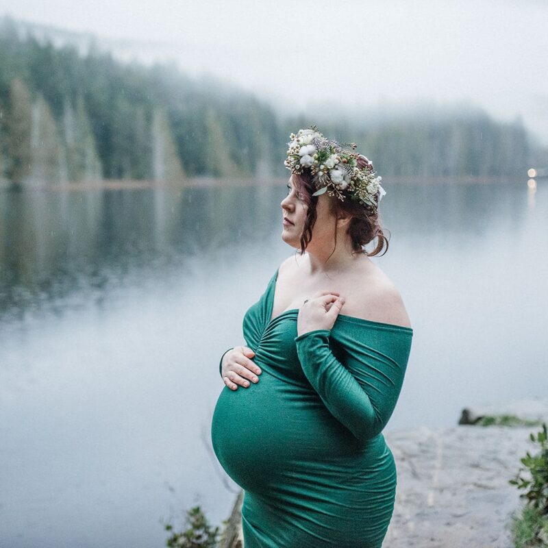 Pregnant woman in an emerald green off-shoulder dress stands in front of a lake caressing her bump during her maternity photoshoot