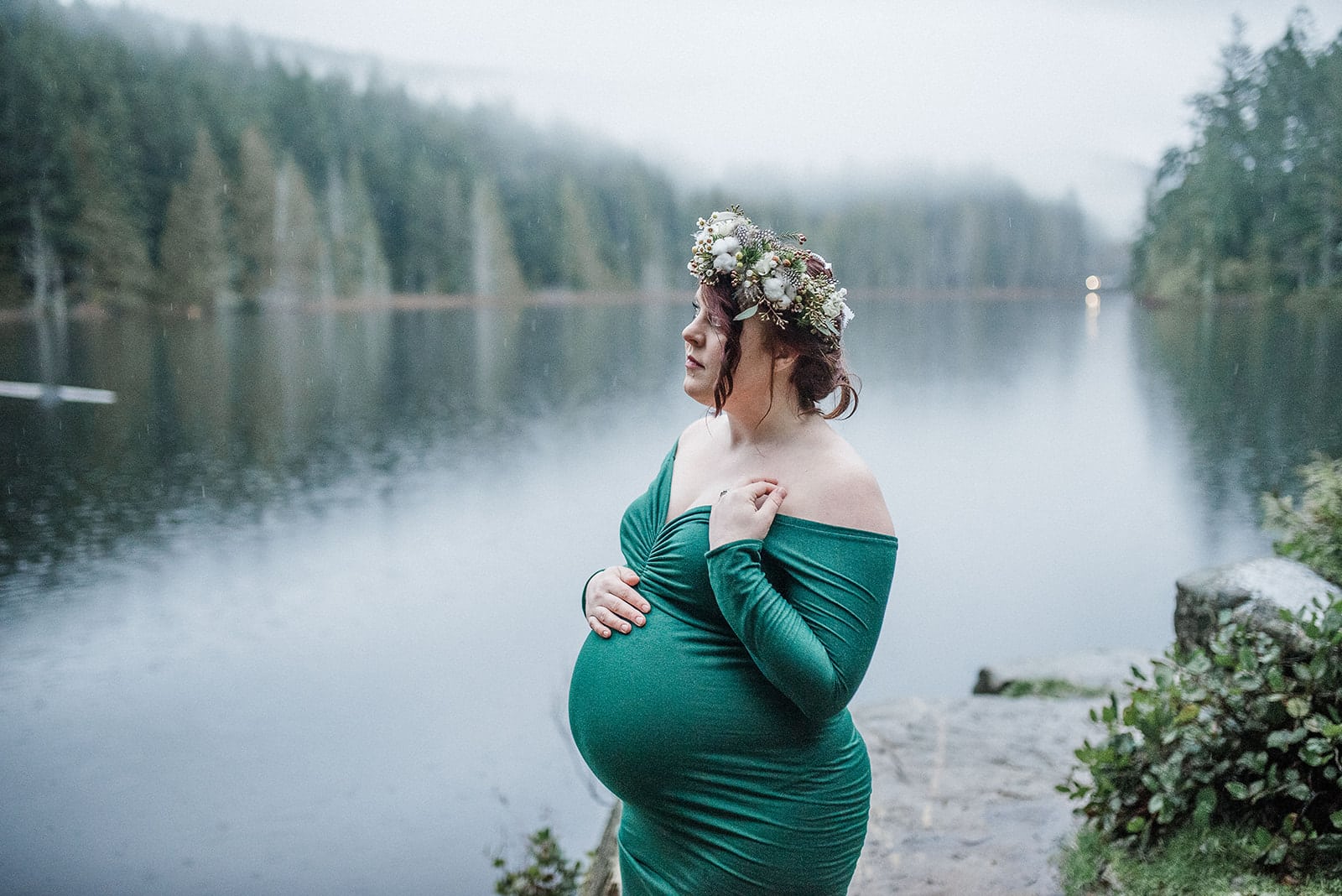 Pregnant woman in an emerald green off-shoulder dress stands in front of a lake caressing her bump during her maternity photoshoot