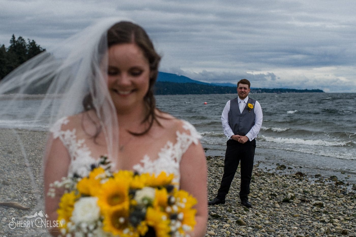 groom stands behind his bride as she smiles at her gorgeous bouquet by Coastal Wedding Events