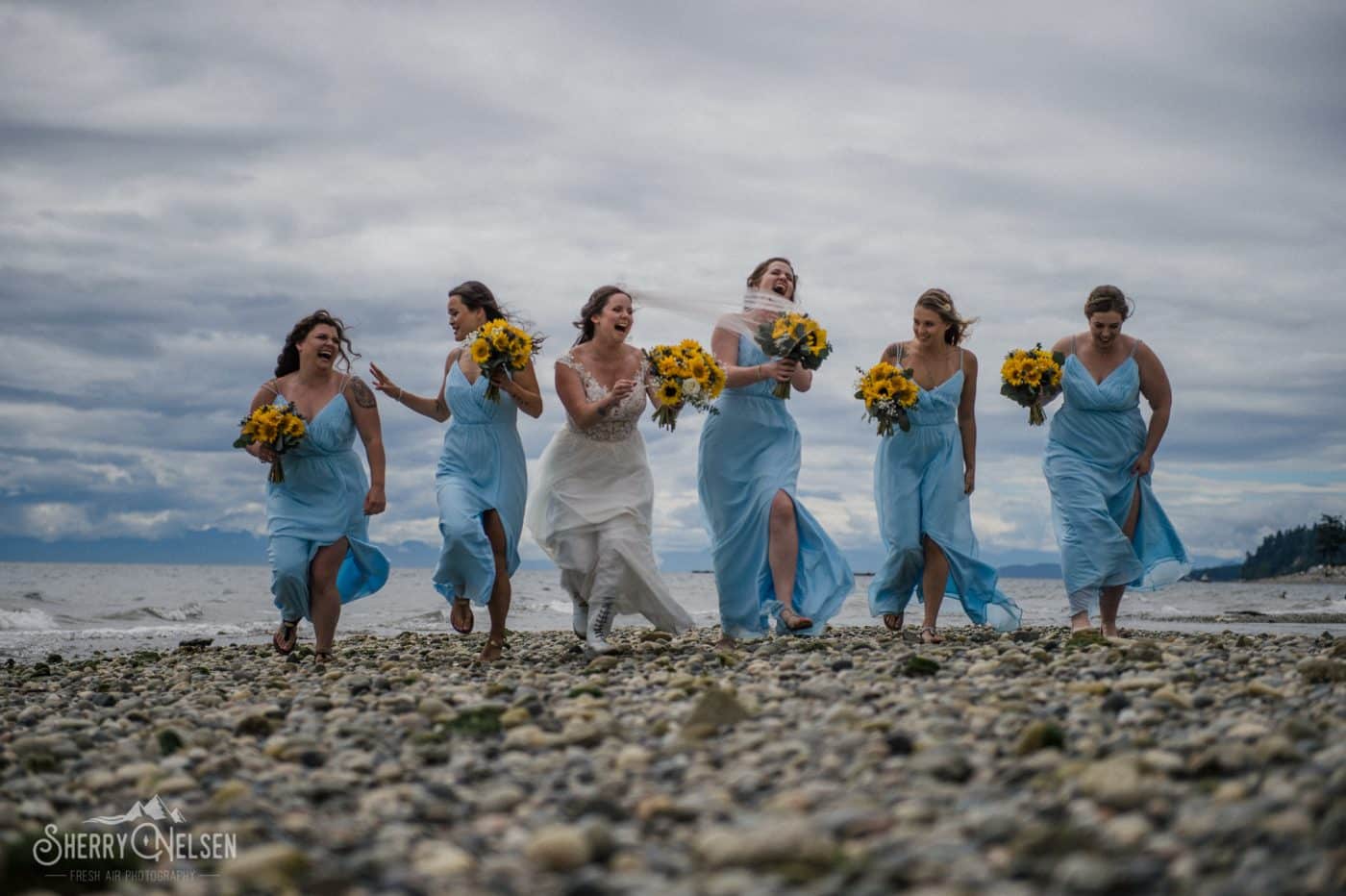 the bride and her bridesmaids run along the beach in happiness as the wind blows their dresses and viels around