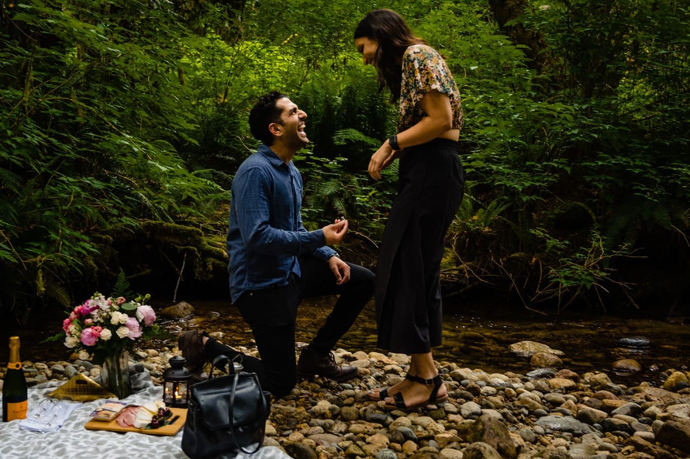 young brown man down on one knee laughs and smiles as he proposes to his beautiful brown girlfriend and she accepts. Images captured by planning and proposal photographer fresh air photography