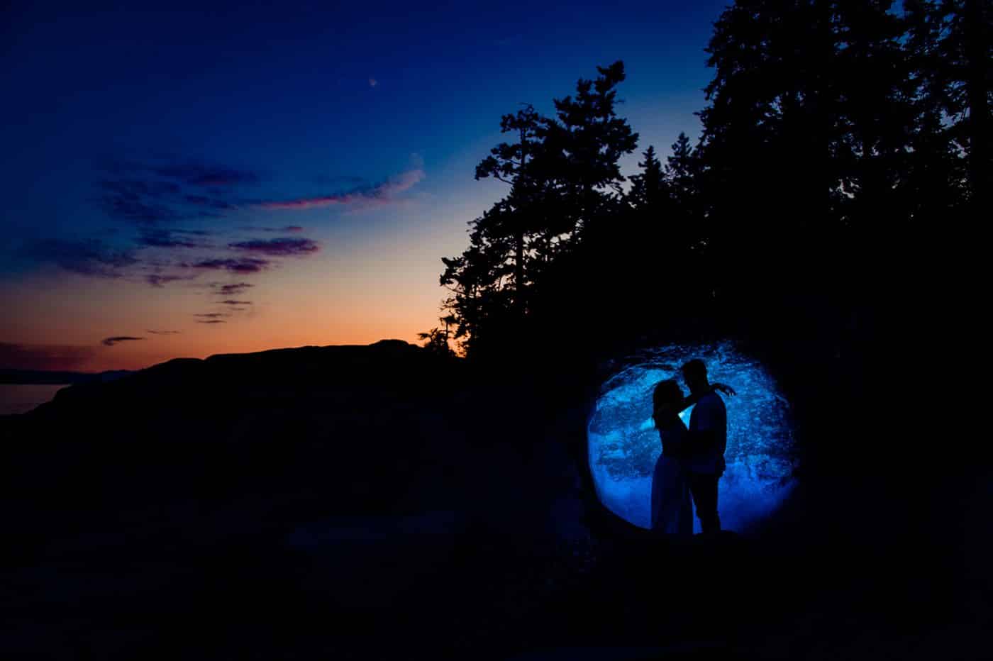 man and woman stand spotlighted in silhouette against a rock with the sun setting toward a blue sky behind them.