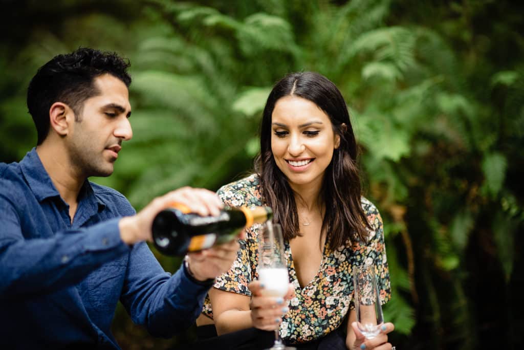 man and woman of middle eastern descent celebrate their engagement in a forest on the Sunshine Coast with glasses of champagne and smiles