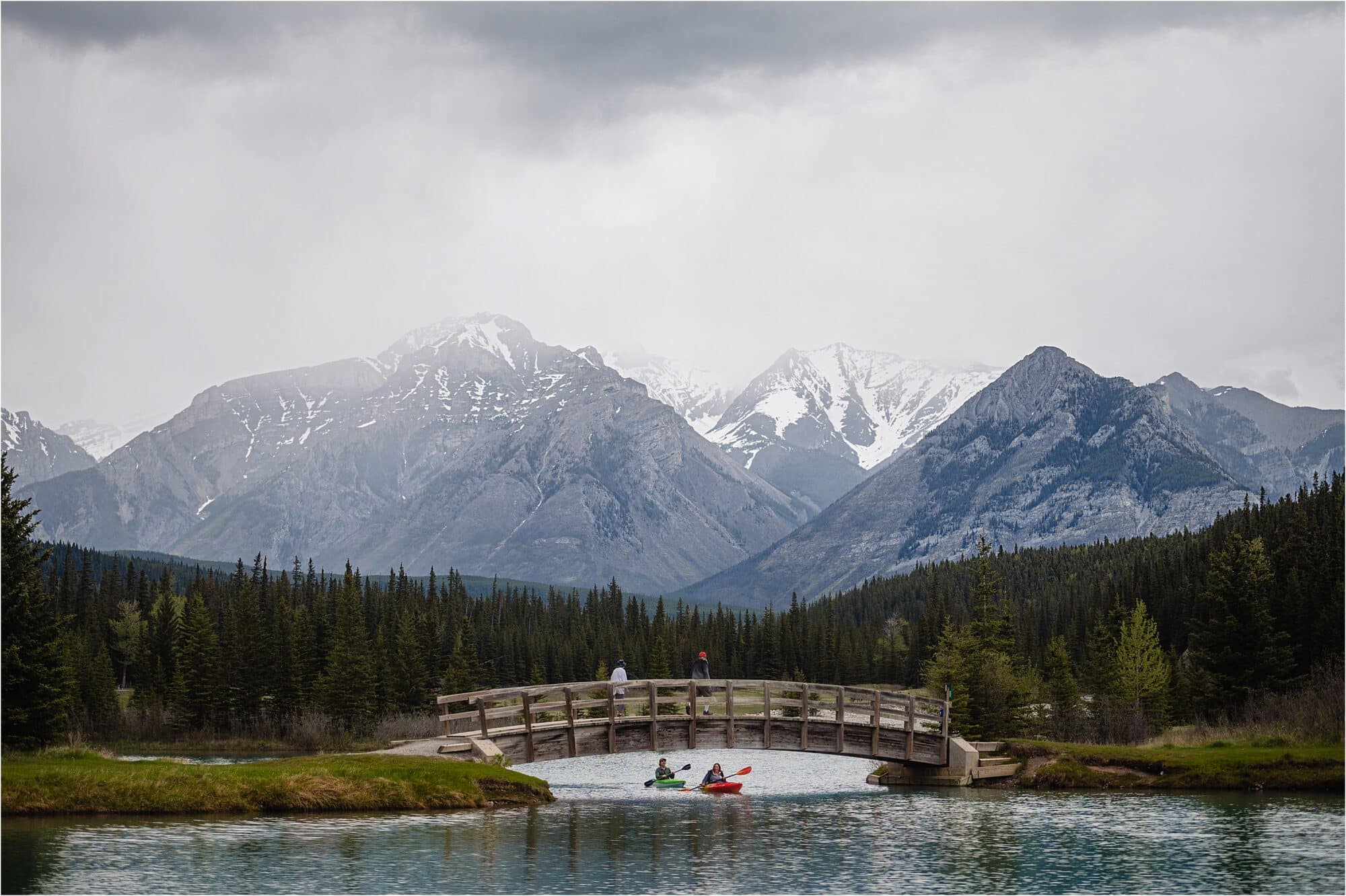 engagement shoot with Fresh Air Photography in the rockies with couple kayaking under a bridge with mountains behind them.