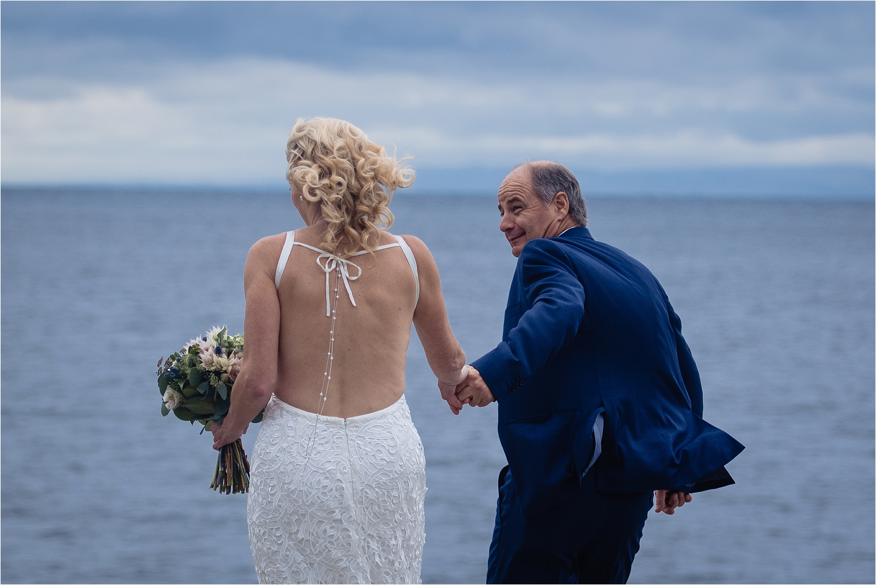 groom leads his bride along the water on a windy day