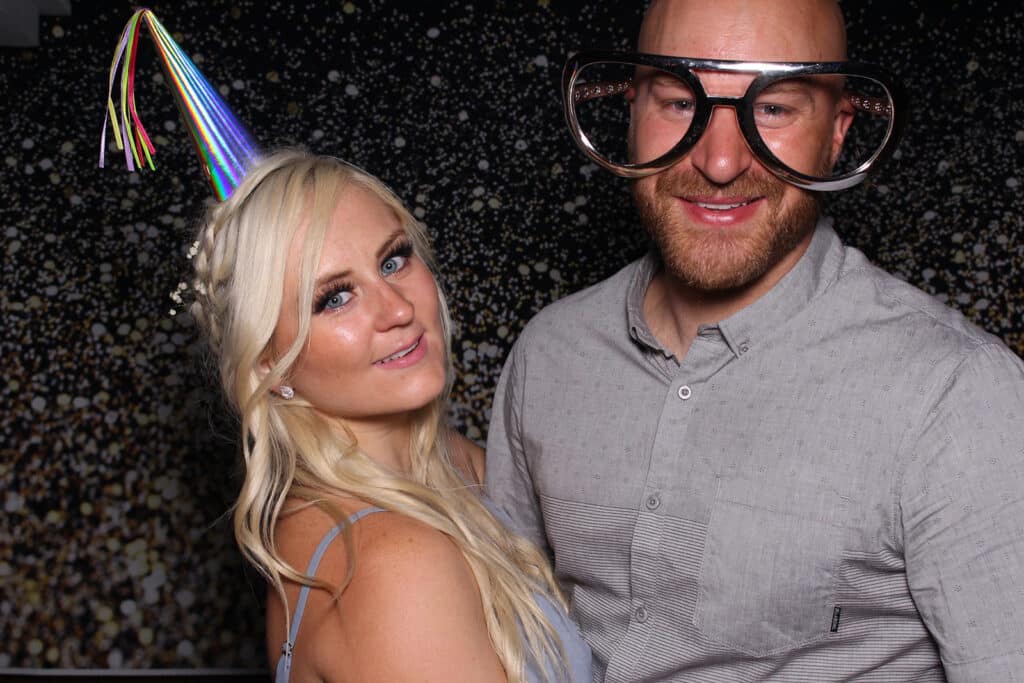 Blonde woman in a party hat smiles at the camera inside a photo booth next to a gentleman in a grey shirt and big glasses