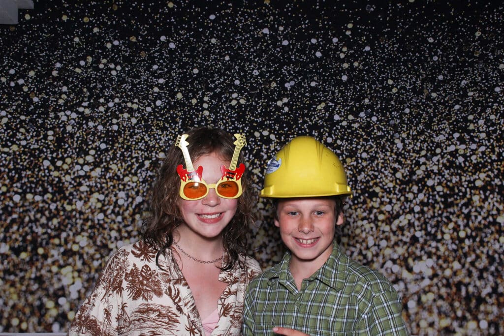 A young woman wearing guitar-shaped sunglasses has her photo taken next to a boy wearing a construction hat at a fun event