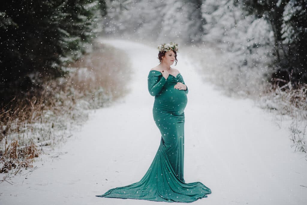 Pregnant woman in a stunning flower crown and floor-length emerald gown stands on a snowy pathway during her winter maternity photoshoot in BC