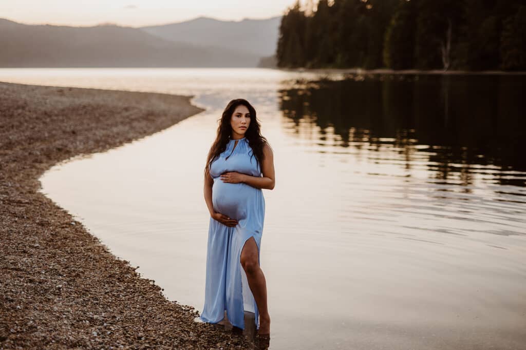 Woman in a blue dress poses with her hands around her baby bump at sunset during her Sunshine Coast maternity session in BC