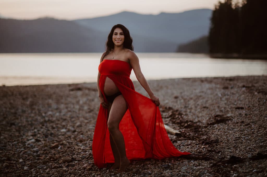 Woman wears a flowy, red outfit that splits up the middle to showcase her gorgeous baby bump on a pebble-lined beach during her pregnancy photoshoot
