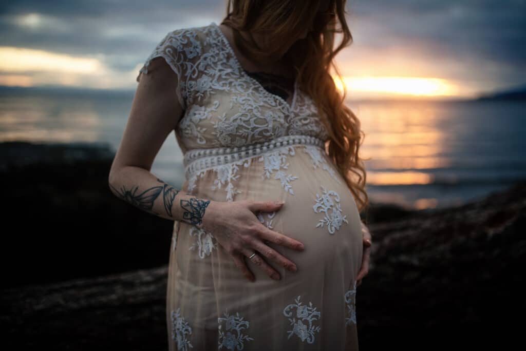 woman with long, blonde hair in a white and nude detailed dress holds her large baby bump at sunset during her maternity photoshoot