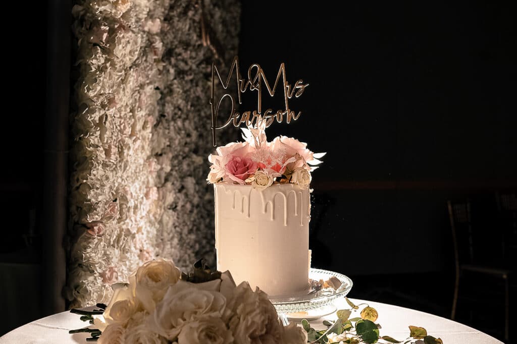 A white wedding cake topped with white and pink roses and a topper that reads "Mr & Mrs Pearson"